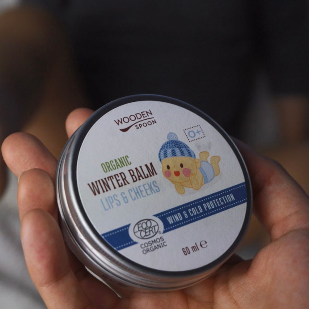 Runde Dose mit Deckel, gehalten in einer Hand. Aufschrift: Wooden Spoon, Organic Winter Balm Lips & Cheeks. Baby-Illustration. Eco Cert Cosmos Organic.