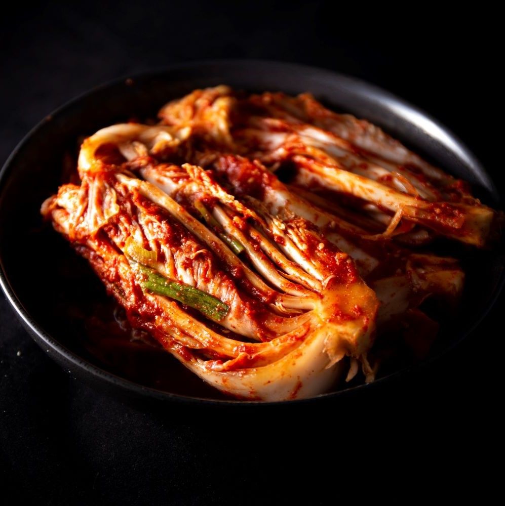 Kimchi in einer schwarzen Schüssel. Das Kimchi ist rot und hat eine blättrige Textur. Grüne Zwiebeln sind sichtbar.