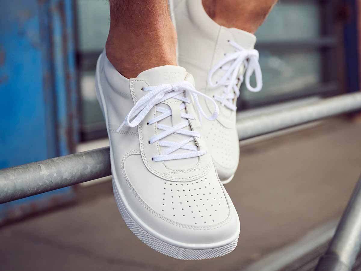 Zwei weiße Lederschnürschuhe. Perforierte Zehenpartie, breite Passform. Schnürsenkel. Auf einer Metallstange platziert.