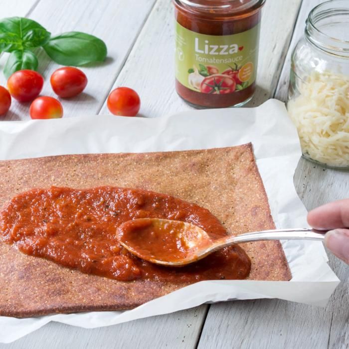 Tomatensauce wird auf einem Teigfladen mit einem Löffel verteilt. Im Hintergrund: Tomaten, Basilikum, Glas mit Käse und Tomatensauce.