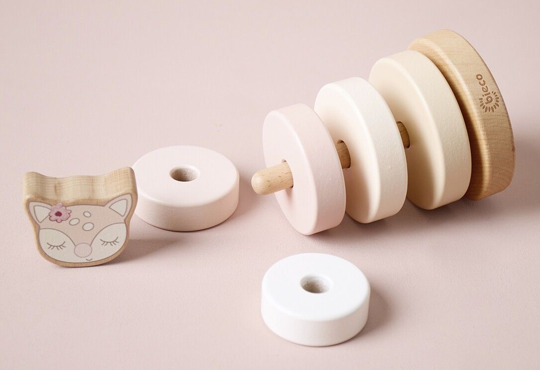 Stapelturm-Teile: Holzsockel, Ringe, Reh-Kopf. Teile liegen. Pastelltöne. Logo auf Sockel. Rosa Hintergrund.