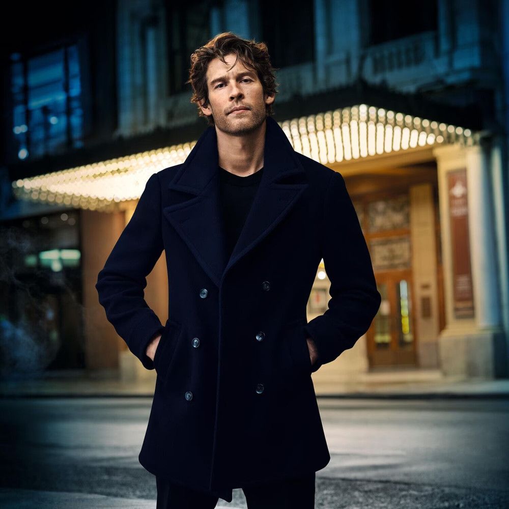 Mann in dunkelblauem Mantel, vor einem Gebäude mit Lichtern. Der Mann steht mit verschränkten Armen.