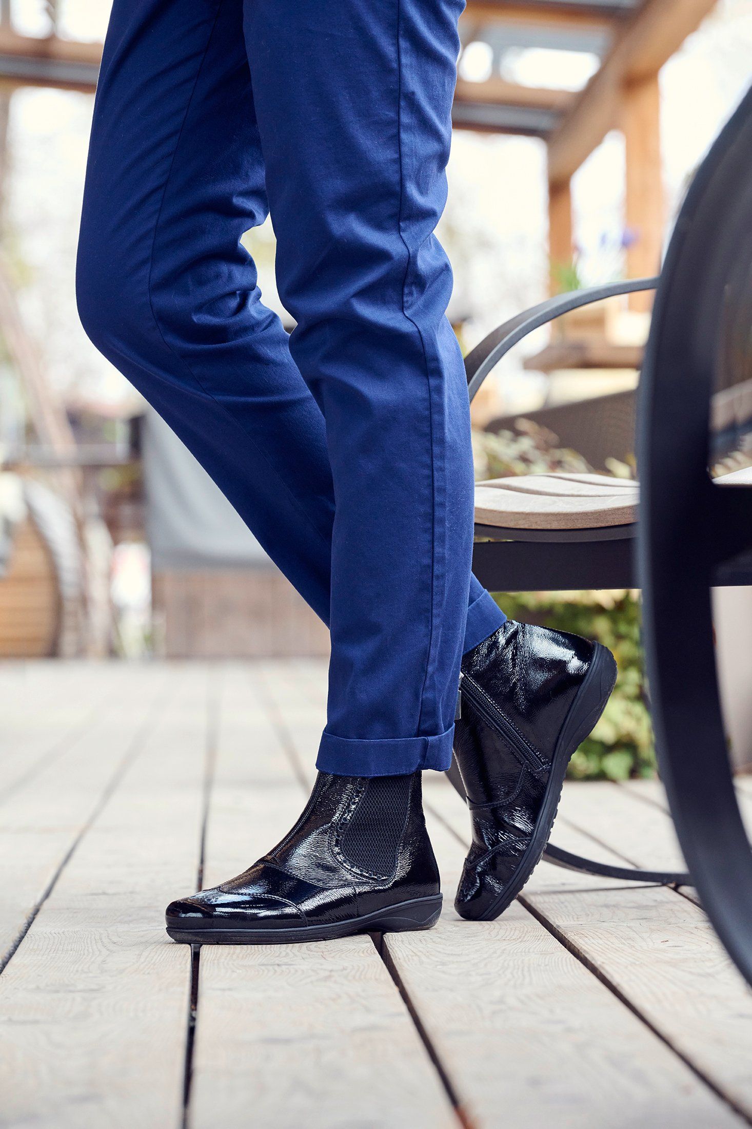 Person trägt blaue Hose und schwarze Stiefeletten. Glänzendes Obermaterial, Reißverschluss, breite Passform.