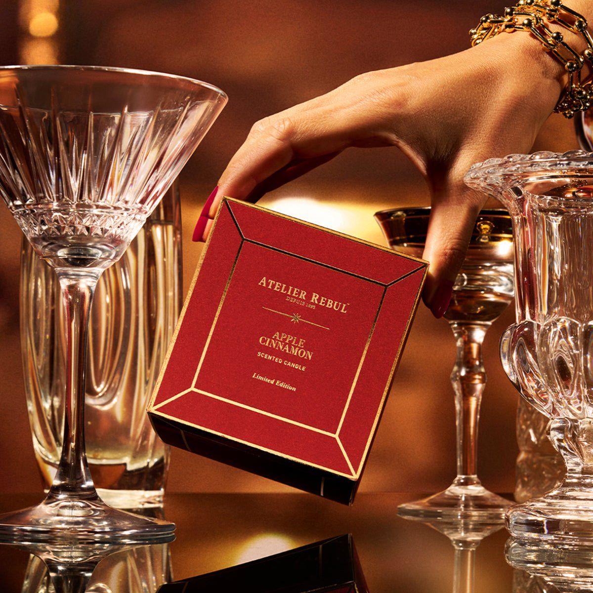 Rote Geschenkbox mit goldener Umrandung. Atelier Rebul-Logo und Produktname. Hände halten die Box. Gläser im Hintergrund.