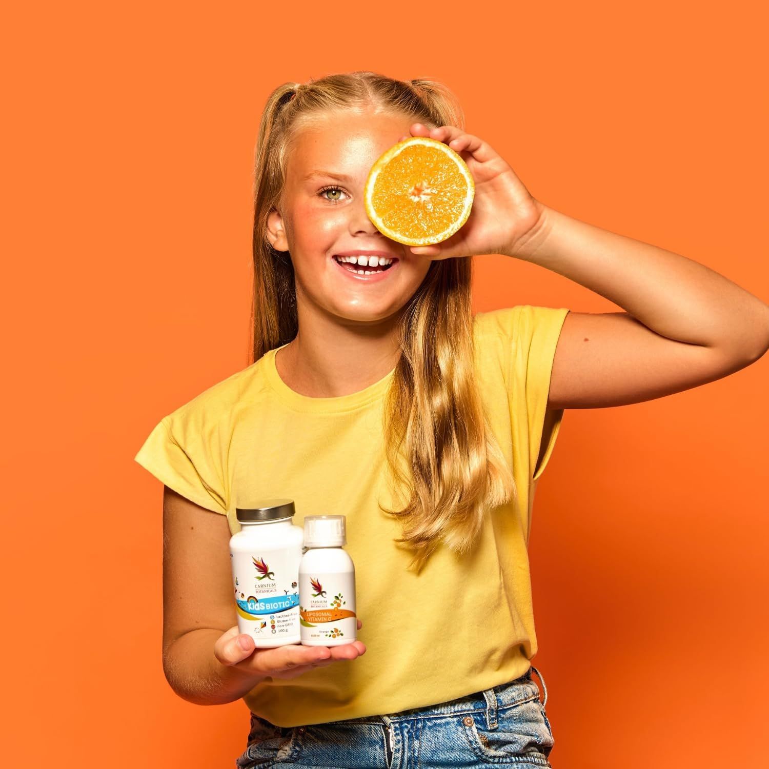 Mädchen hält eine Orange vor einem orangefarbenen Hintergrund. Zwei Flaschen mit Aufschrift Kids Biotic.