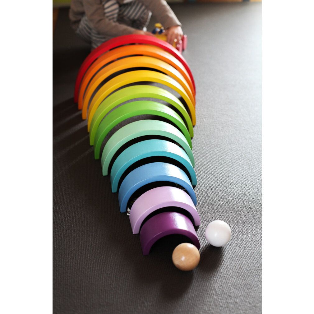 Kind spielt mit Regenbogen-Holzbausteinen. Die Steine sind in einer Reihe angeordnet. Zwei Kugeln liegen daneben.