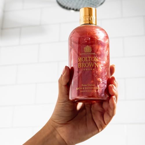 Hand hält eine Flasche Molton Brown Rose Dunes Duschgel. Hintergrund: weiß geflieste Wand und Duschkopf.