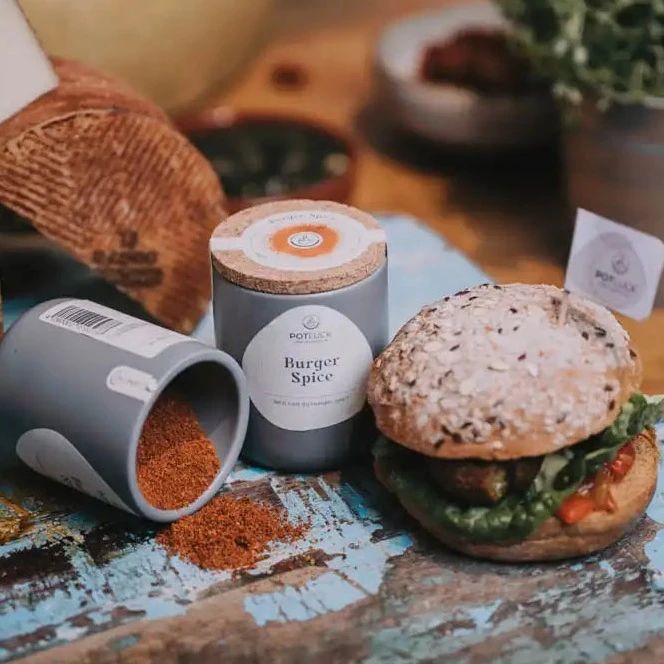 Graue Dose POTLUCK Burger Spice, daneben umgekippte Dose mit rotem Inhalt. Burger mit Sesambrötchen. Auf Holzuntergrund.
