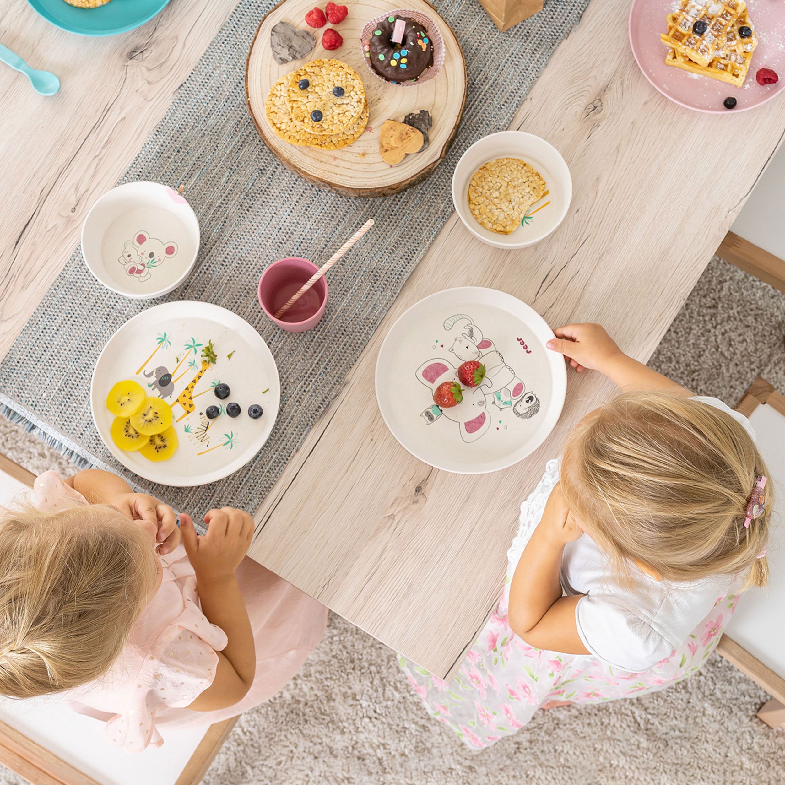 Zwei Mädchen sitzen an einem Tisch mit Kindergeschirr. Teller mit Wildtier-Design, Waffeln und Kuchen. Rosa und blaue Becher.