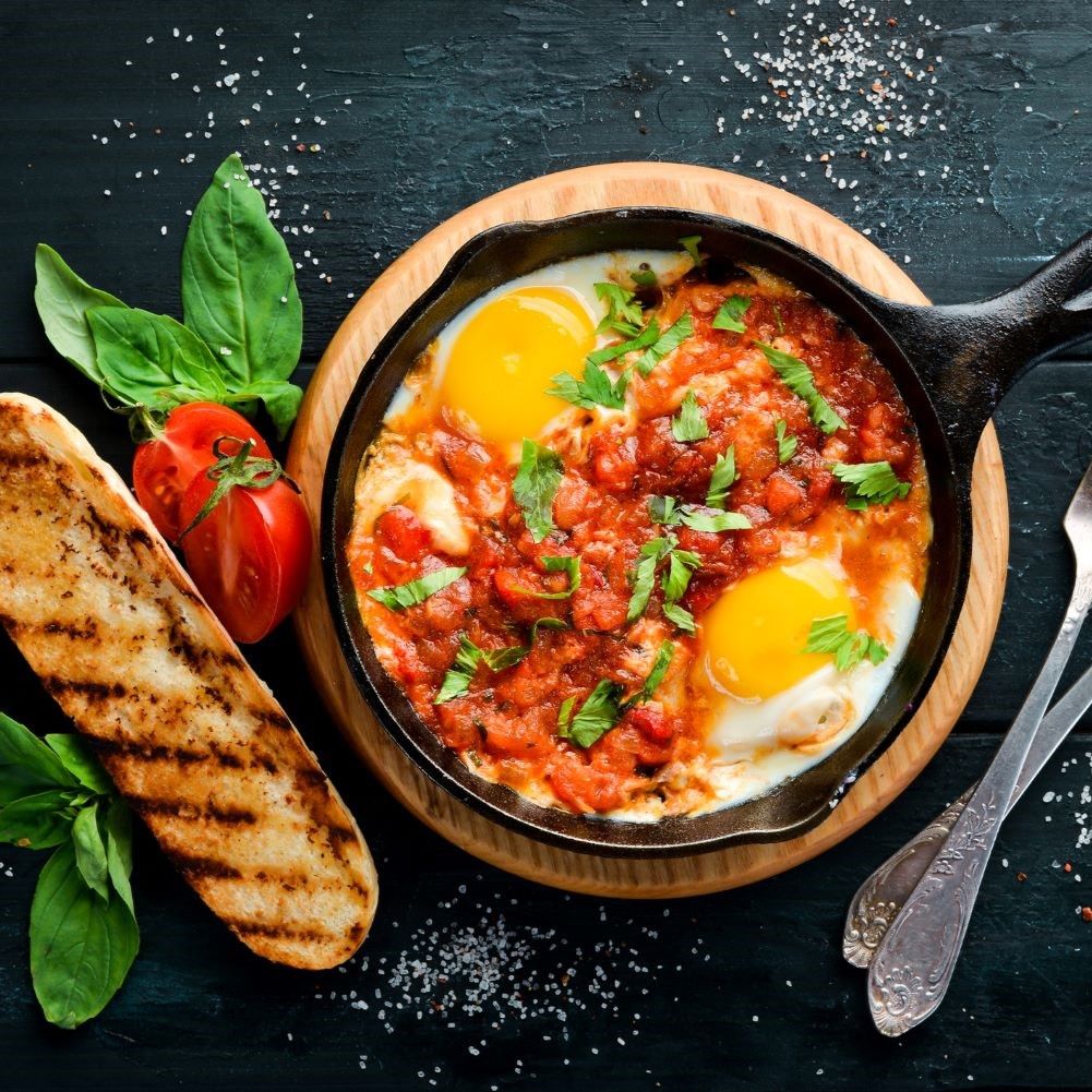 Shakshuka in Pfanne mit Eiern, Tomaten, Kräutern. Auf Holzbrett mit Brot, Tomate und Besteck. Schwarzer Hintergrund.