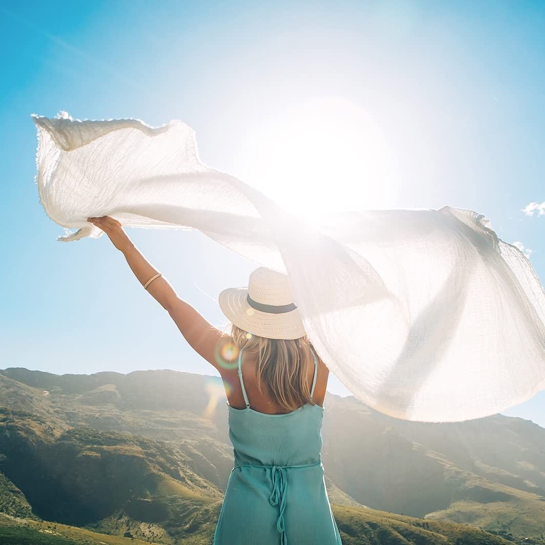 Donna con cappello e tessuto bianco davanti a un paesaggio montuoso. Tessuto al vento, sole sullo sfondo.