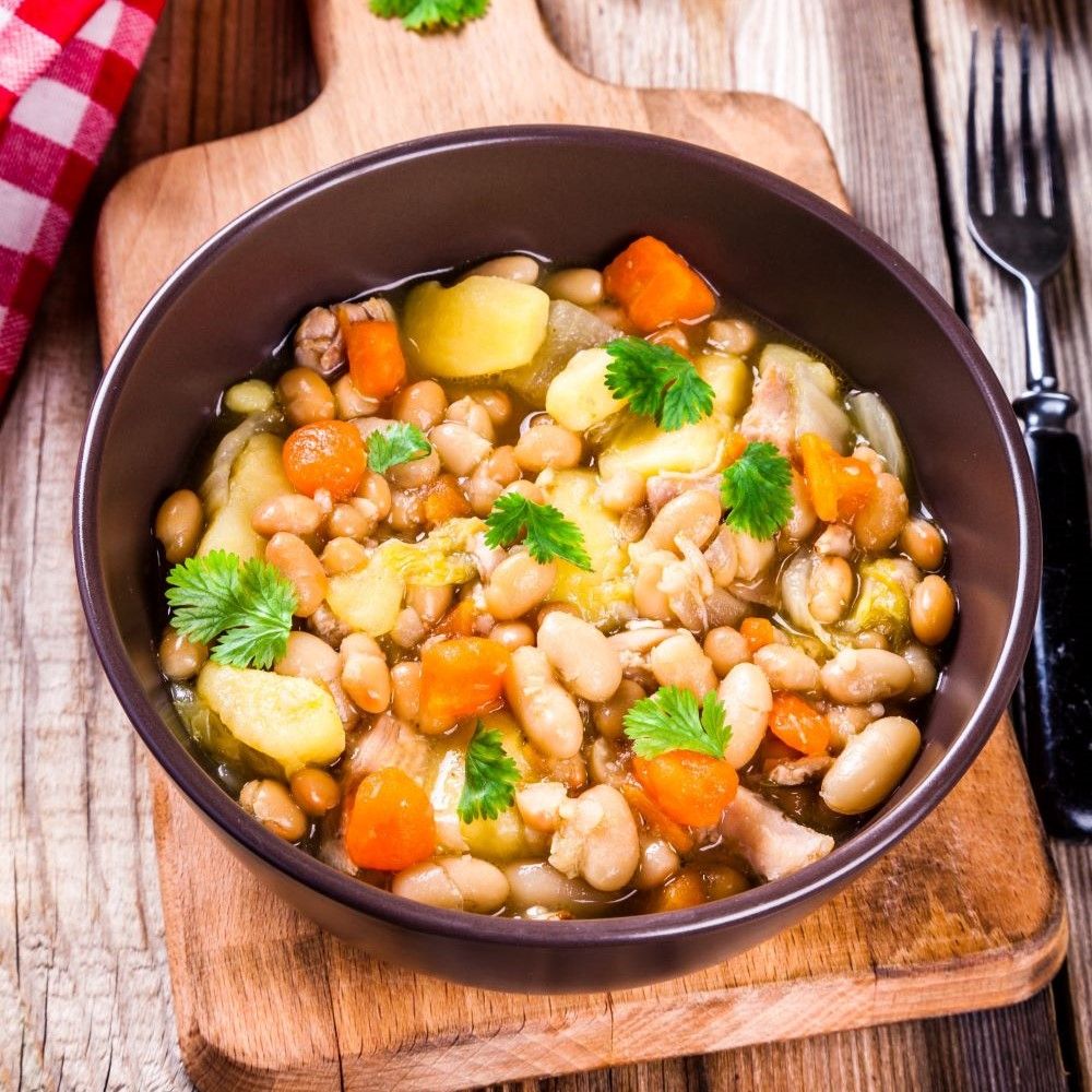 Braune Schüssel mit Eintopf. Enthält Bohnen, Kartoffeln, Karotten und Kräuter. Auf einem Holzbrett.