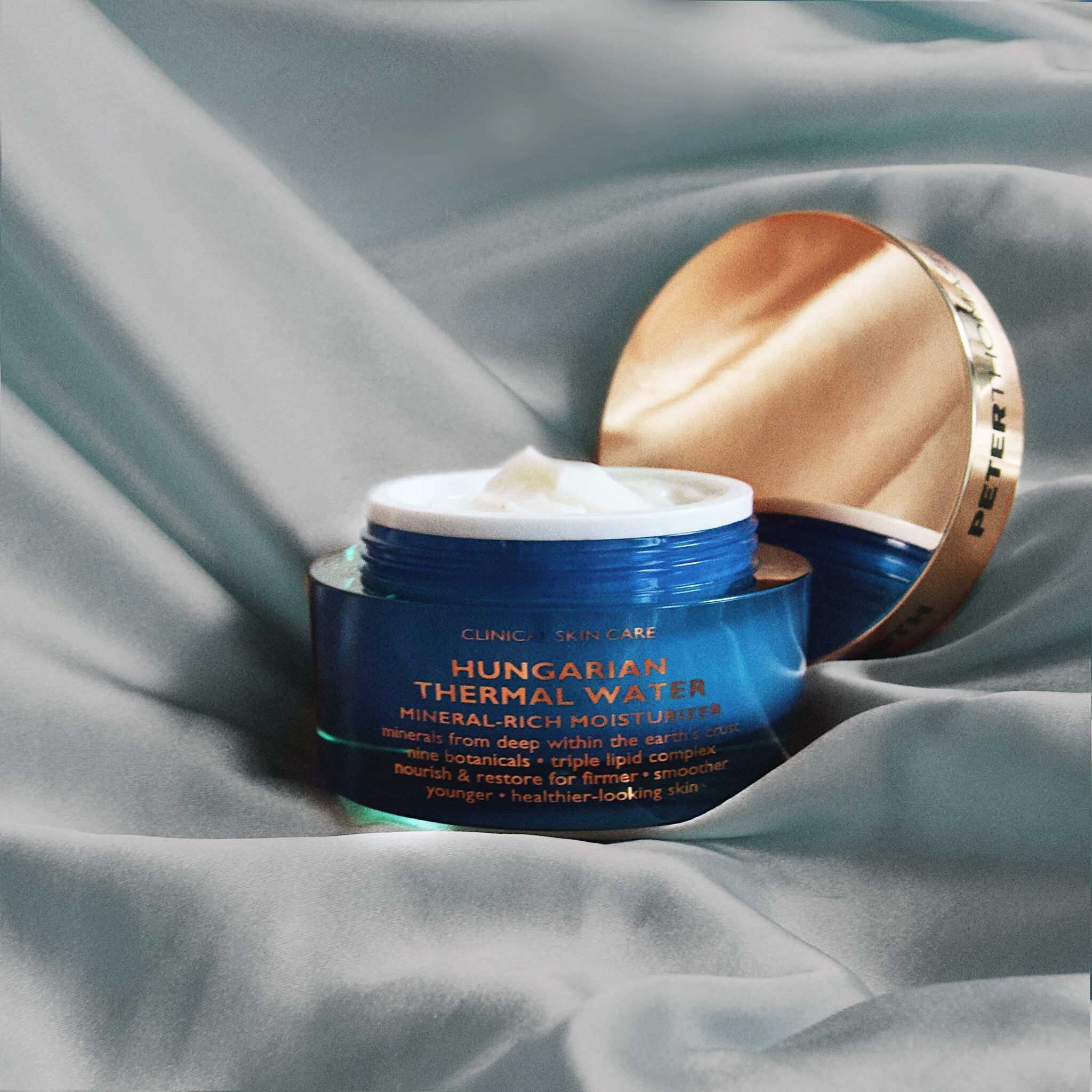 Geöffneter Tiegel mit goldfarbenem Deckel auf hellblauem Stoff. Produktname und Text sichtbar. Creme im Inneren.