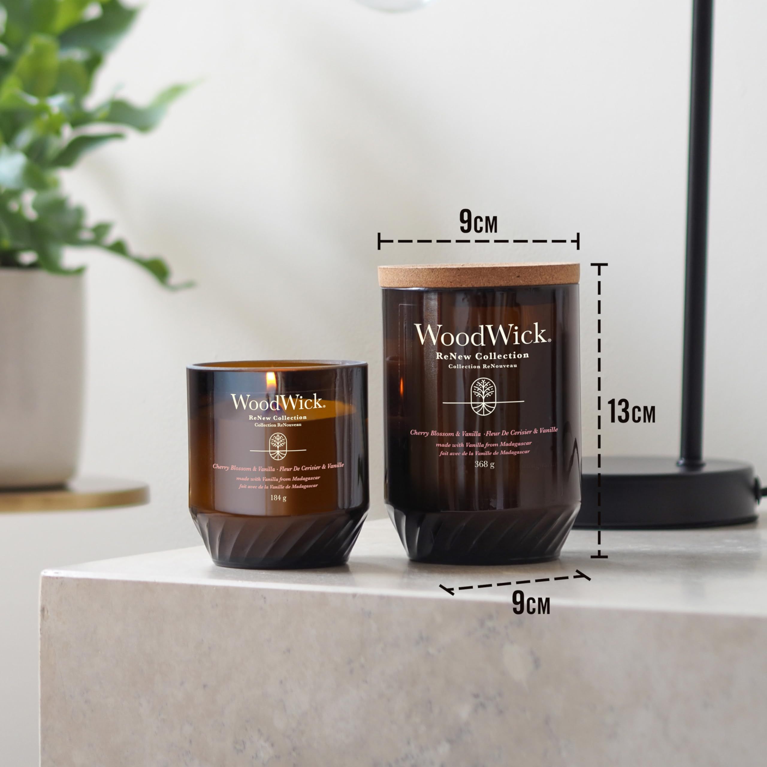 Zwei Kerzen mit Korkdeckeln. Aufschrift: WoodWick. Maße: 9 cm x 13 cm. Eine Kerze brennt. Auf Tisch platziert.