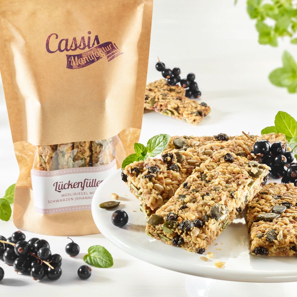 Müsli-Riegel auf Teller, daneben Beutel. Johannisbeeren und Minze als Dekoration. Logo "Cassis Manufaktur" auf dem Beutel.
