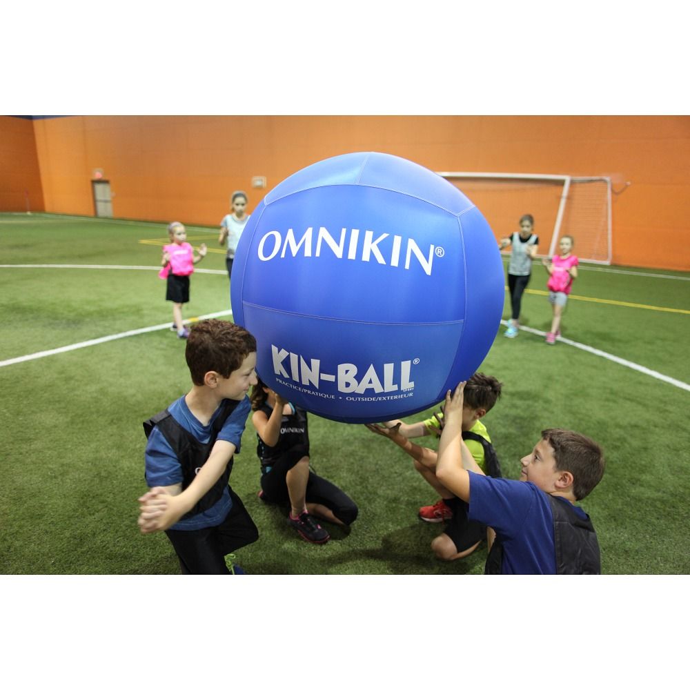 Blauer Omnikin Kin-Ball. Kinder heben den Ball. Aufschrift: OMNIKIN, KIN-BALL. Kinder tragen Westen. Sportplatz im Hintergrund.