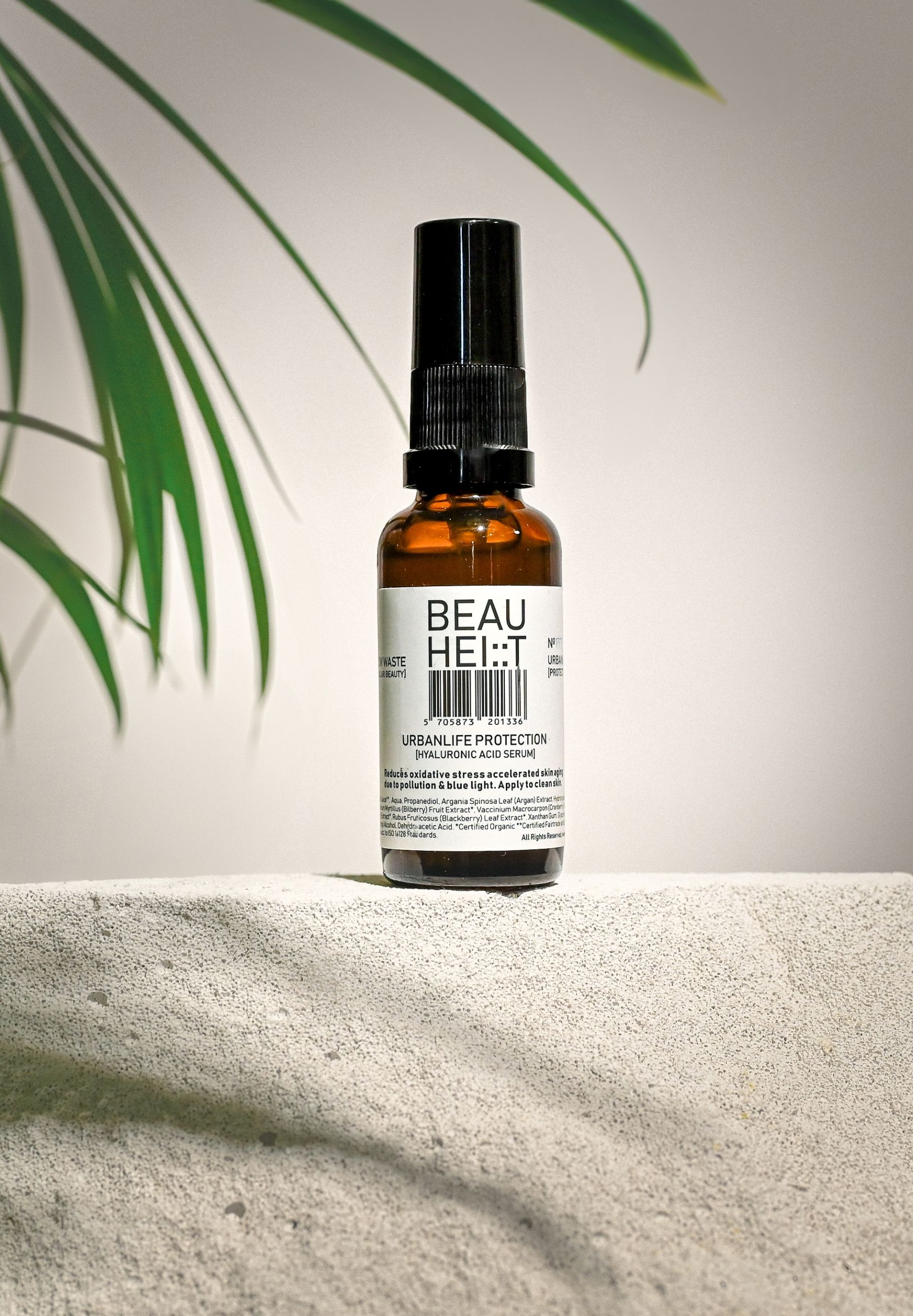 Braune Glasflasche mit schwarzem Sprühkopf. Etikett mit Produktnamen. BEAUHEIT Urbanlife Protection Feuchtigkeitsserum. Auf Stein platziert.