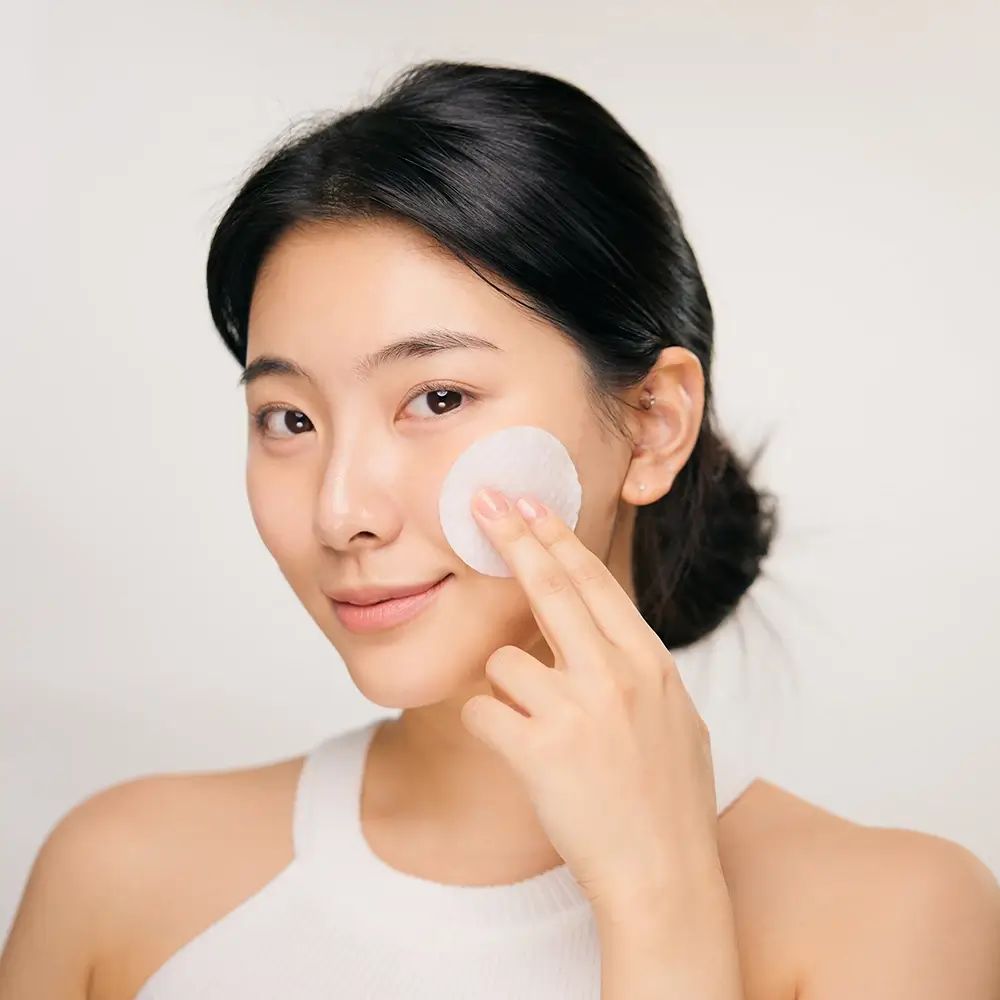 Junge Frau, die mit einem Wattepad ihr Gesicht reinigt. Sie trägt ein weißes Top. Helle Haut, dunkle Haare.