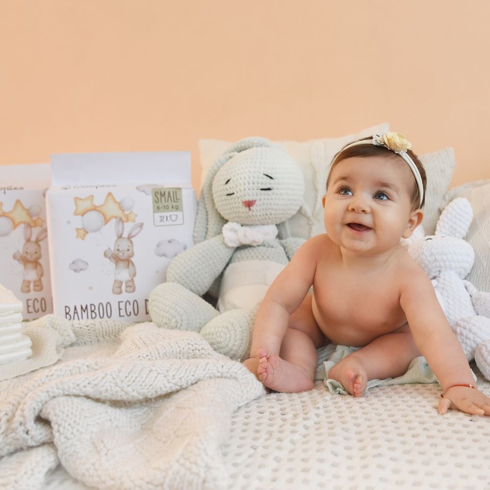 Baby sitzt auf Bett mit Spielzeug. Im Hintergrund: Packung Babywindeln. Aufdruck: Hase mit Luftballons, Sterne, Wolken. Text: Bamboo Eco Diapers, SMALL.