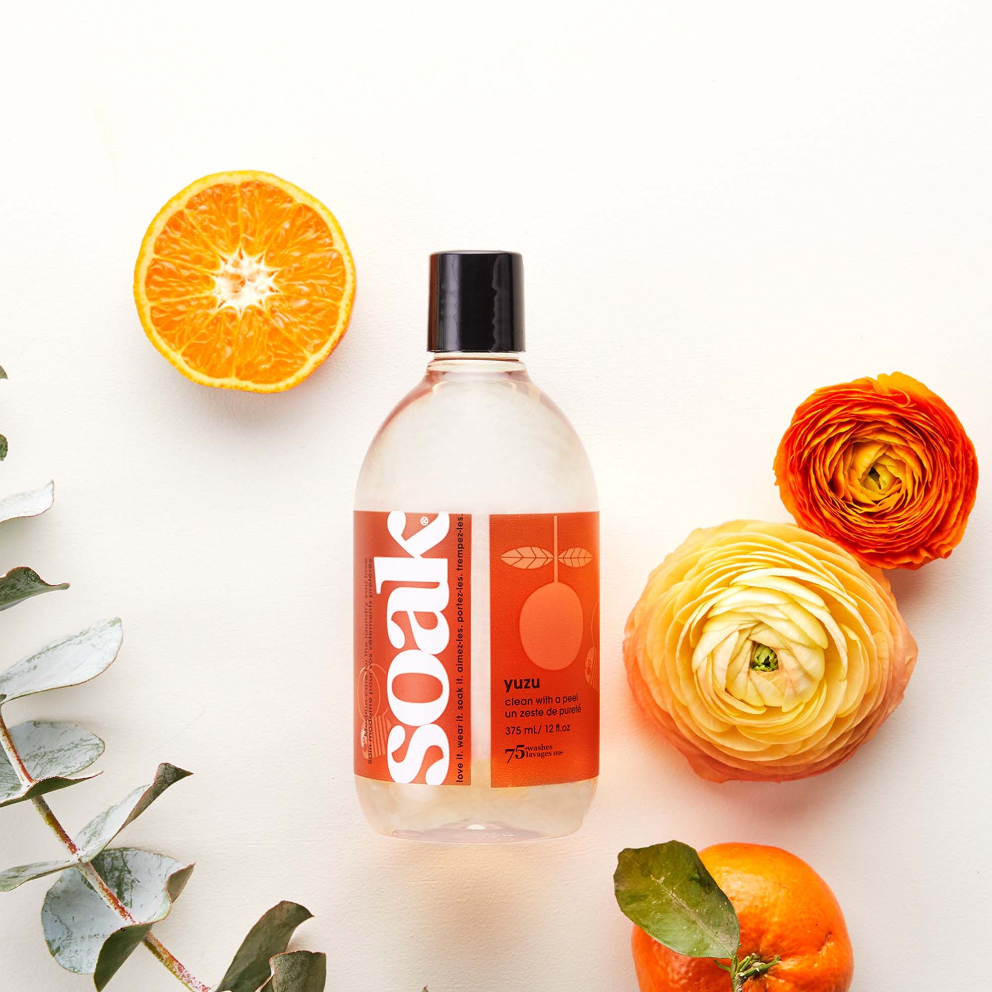 Klare Flasche mit rotem Etikett, "soak"-Logo. Orangen und Blüten auf weißem Hintergrund.