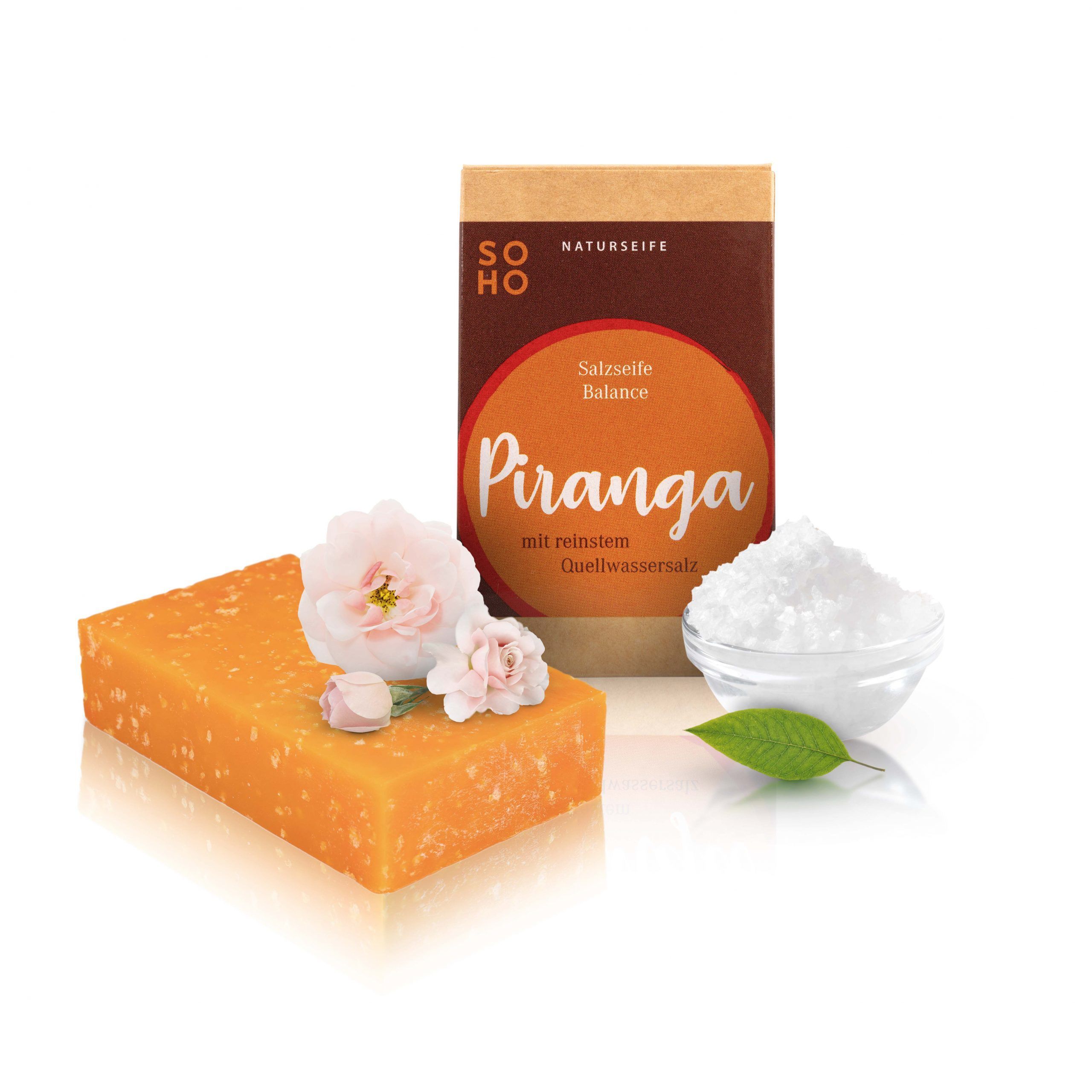 Orangefarbene Salzseife, daneben Verpackung mit "PIRANGA" Schriftzug. Eine Schale mit Salz und Blüten.