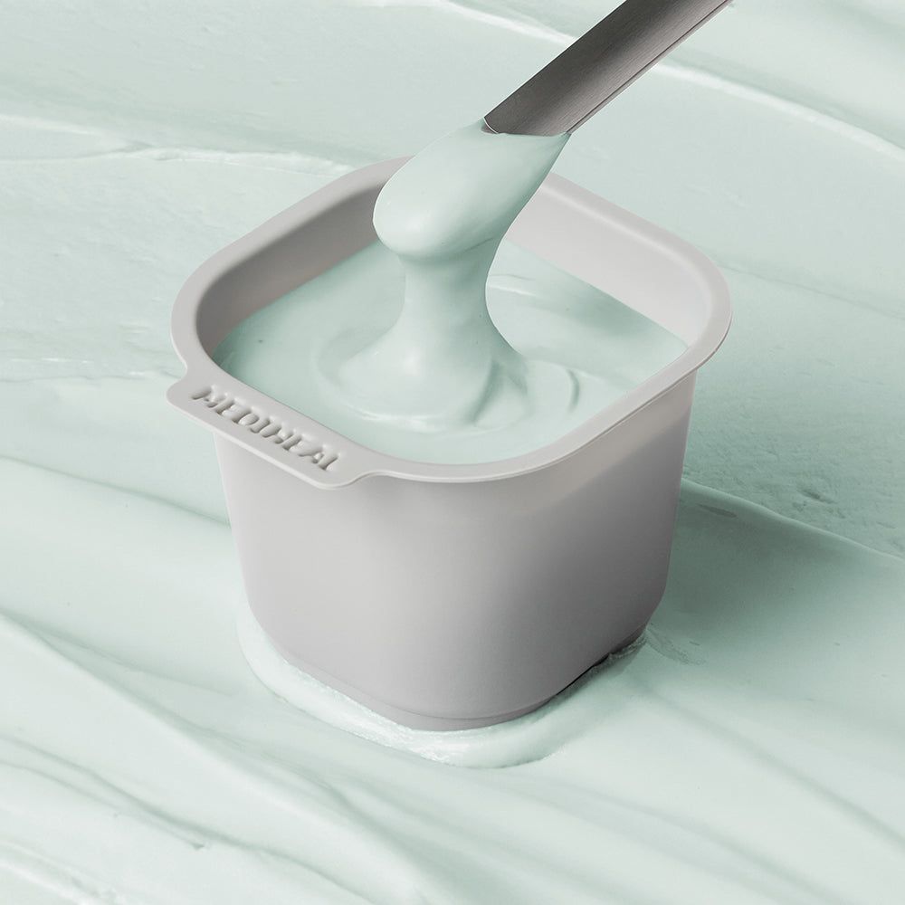 Ein grauer Behälter mit hellgrüner Creme, die von einem Spatel entnommen wird.