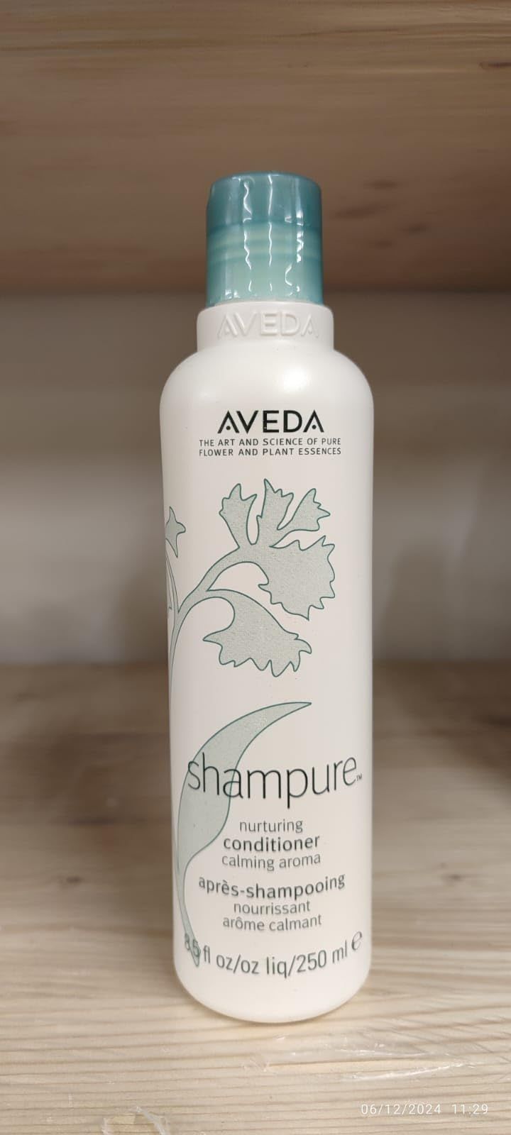 Weiße Flasche mit blauem Deckel. Aufschrift: Aveda, Shampure, Après-shampooing Nourrissant. Floral-Dekor. Auf Regal.