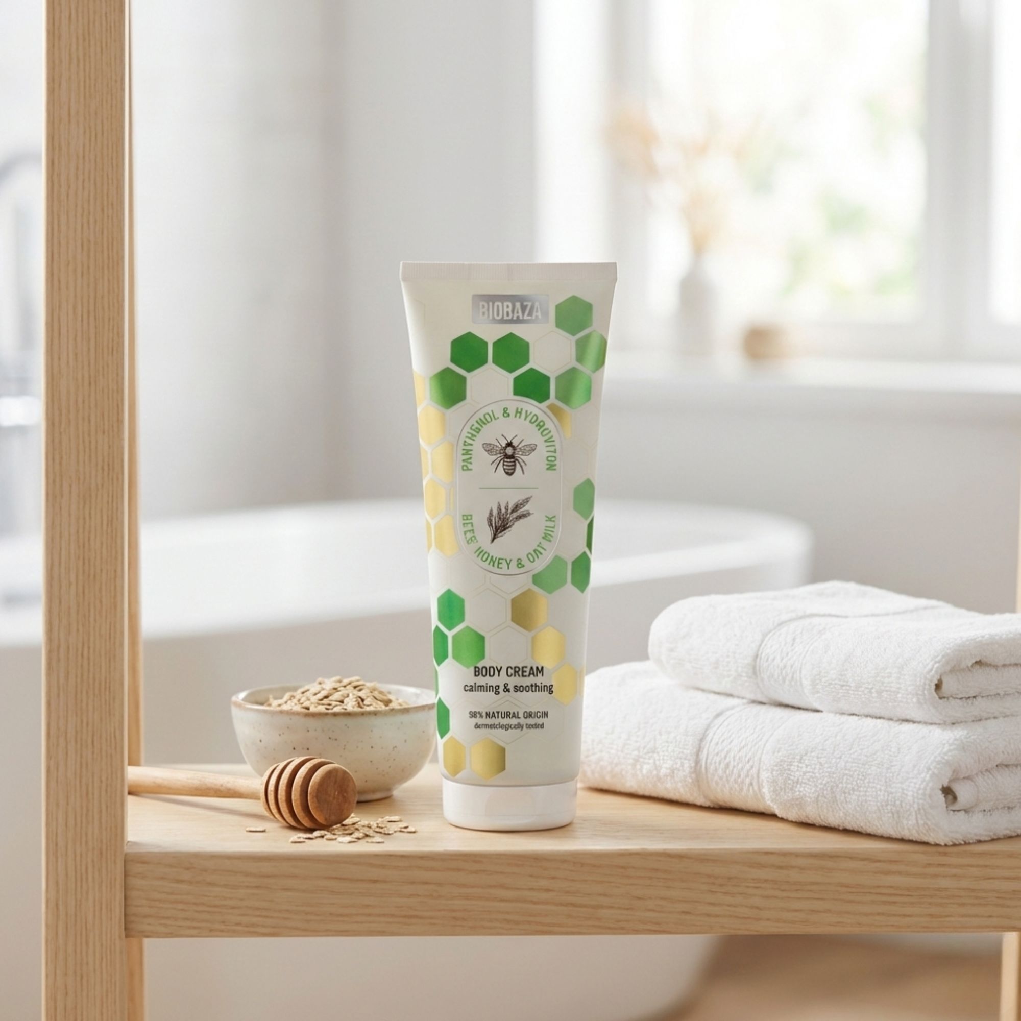 Creme-Tube mit Honig, Hafer und Bienen-Motiv. Auf Regal neben Handtüchern und Schale mit Haferflocken. Natürliche Inhaltsstoffe.