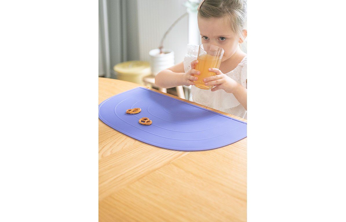 Kind sitzt am Tisch, trinkt. Lila Platzdeckchen mit Brezeln. Halbkreisform mit eingeprägten Bögen.