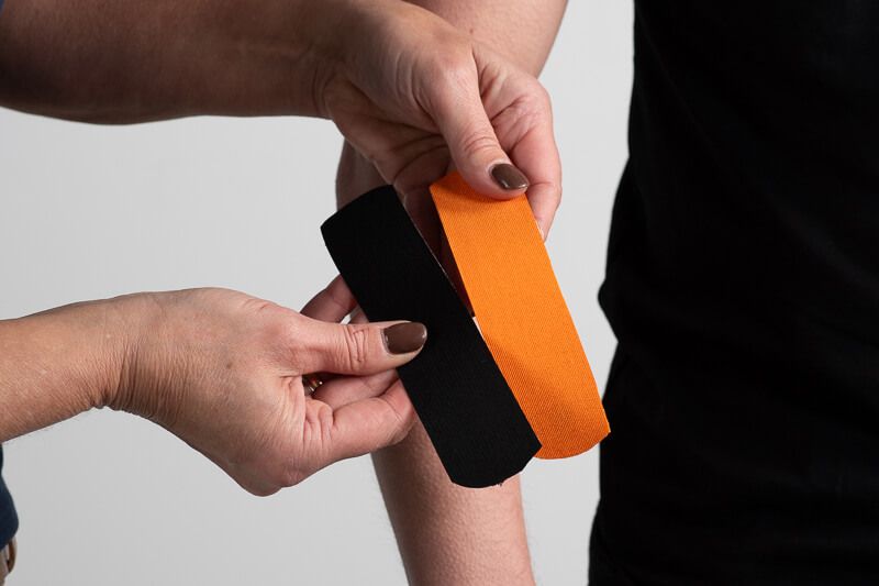 Hände halten zwei Streifen Kinesiotape, orange und schwarz. Der Arm einer Person ist im Hintergrund zu sehen.