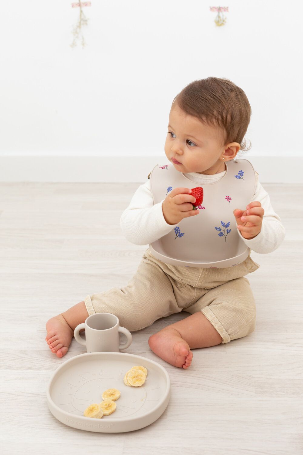 Baby sitzt mit Lätzchen und Teller. Auf dem Teller Bananenscheiben. Baby hält Erdbeere.