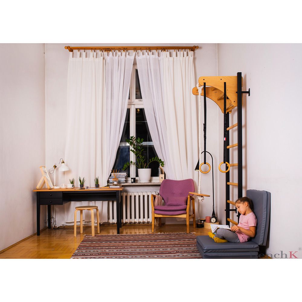 Sprossenwand mit Turnzubehör in einem Raum. Schwarze Sprossen, Holz- und Metallteile. Kind sitzt auf dem Boden.