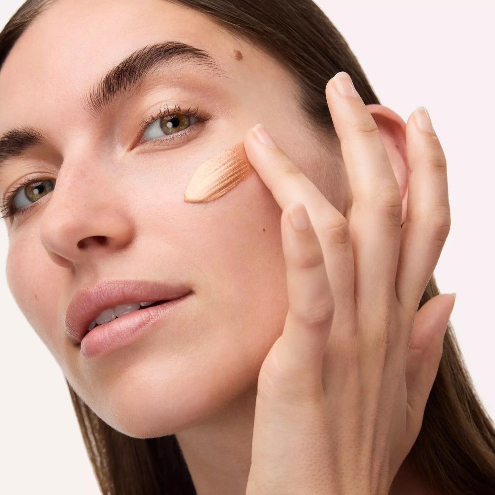 Frau trägt Catrice Bronze Glow Grip Primer auf. Bronzefarbene Flüssigkeit auf dem Finger. Gesicht mit aufgetragener Flüssigkeit.