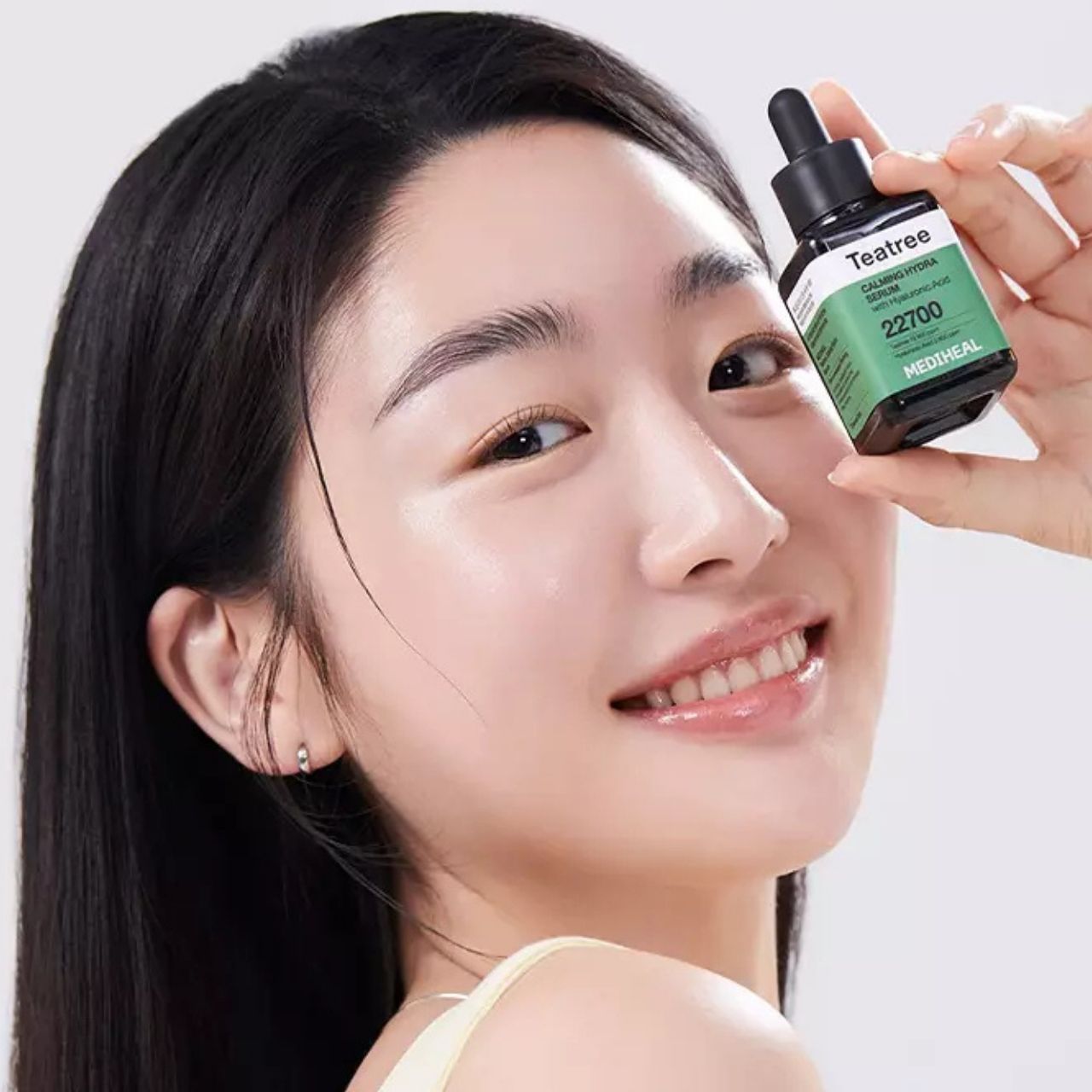 Frau hält MEDIHEAL Teebaumöl Beruhigendes Hydra-Serum Flasche neben ihrem Gesicht.