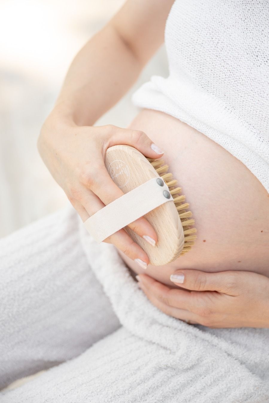 Person hält eine Trockenbürste auf einem Schwangerschaftsbauch. Die Bürste hat einen Holzgriff und helle Borsten.