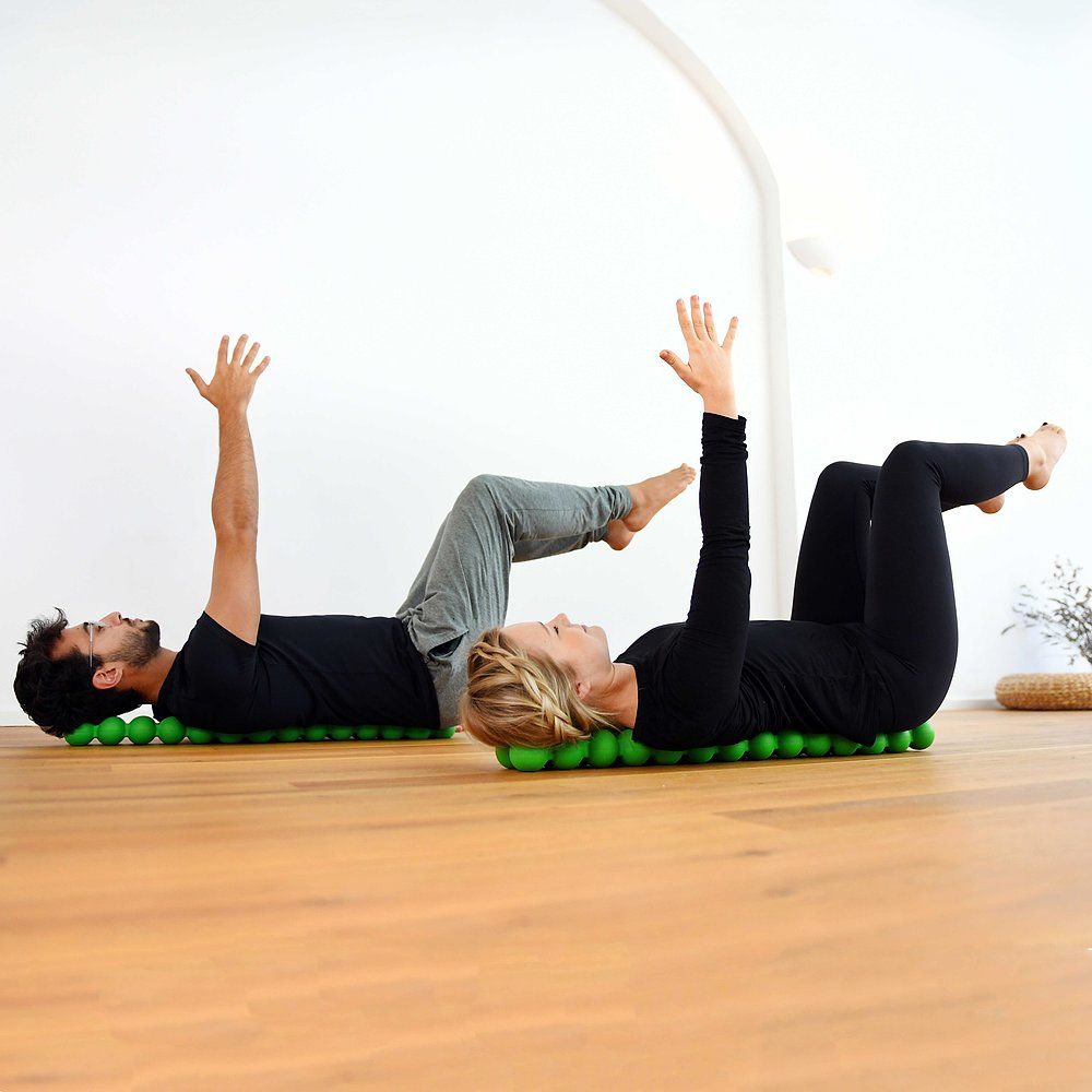 Zwei Personen liegen auf grünen Spinefittern. Arme nach oben gestreckt. Holzfußboden.