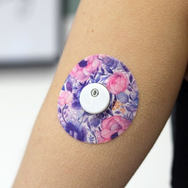 Arm mit angebrachtem Fixierungspflaster. Lila Blumenmuster. Weißes Gerät in der Mitte. Produktname nicht sichtbar.