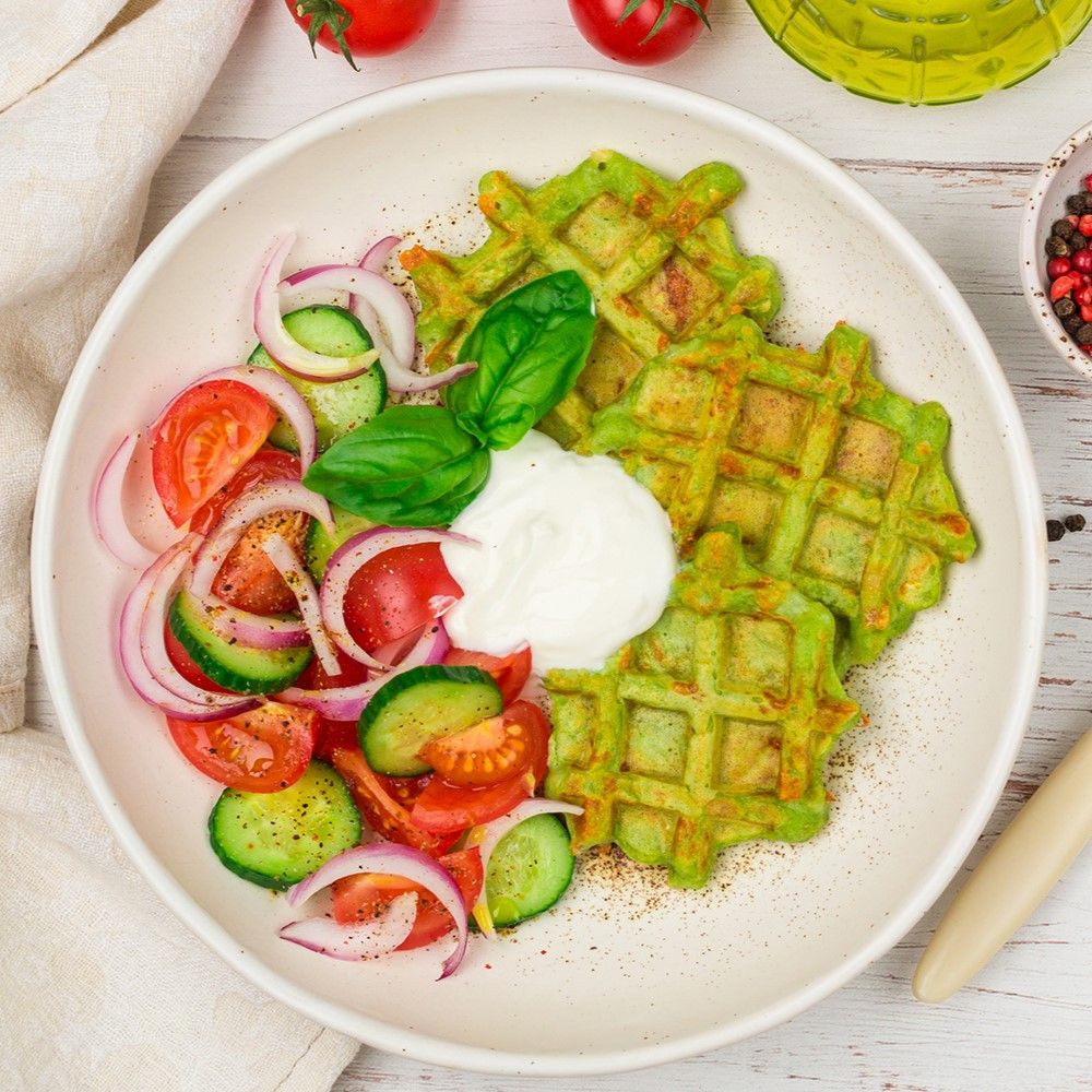 Teller mit grünen Waffeln, Salat aus Tomaten, Gurken und Zwiebeln, sowie einem Klecks weißer Creme. Auf hellem Untergrund.
