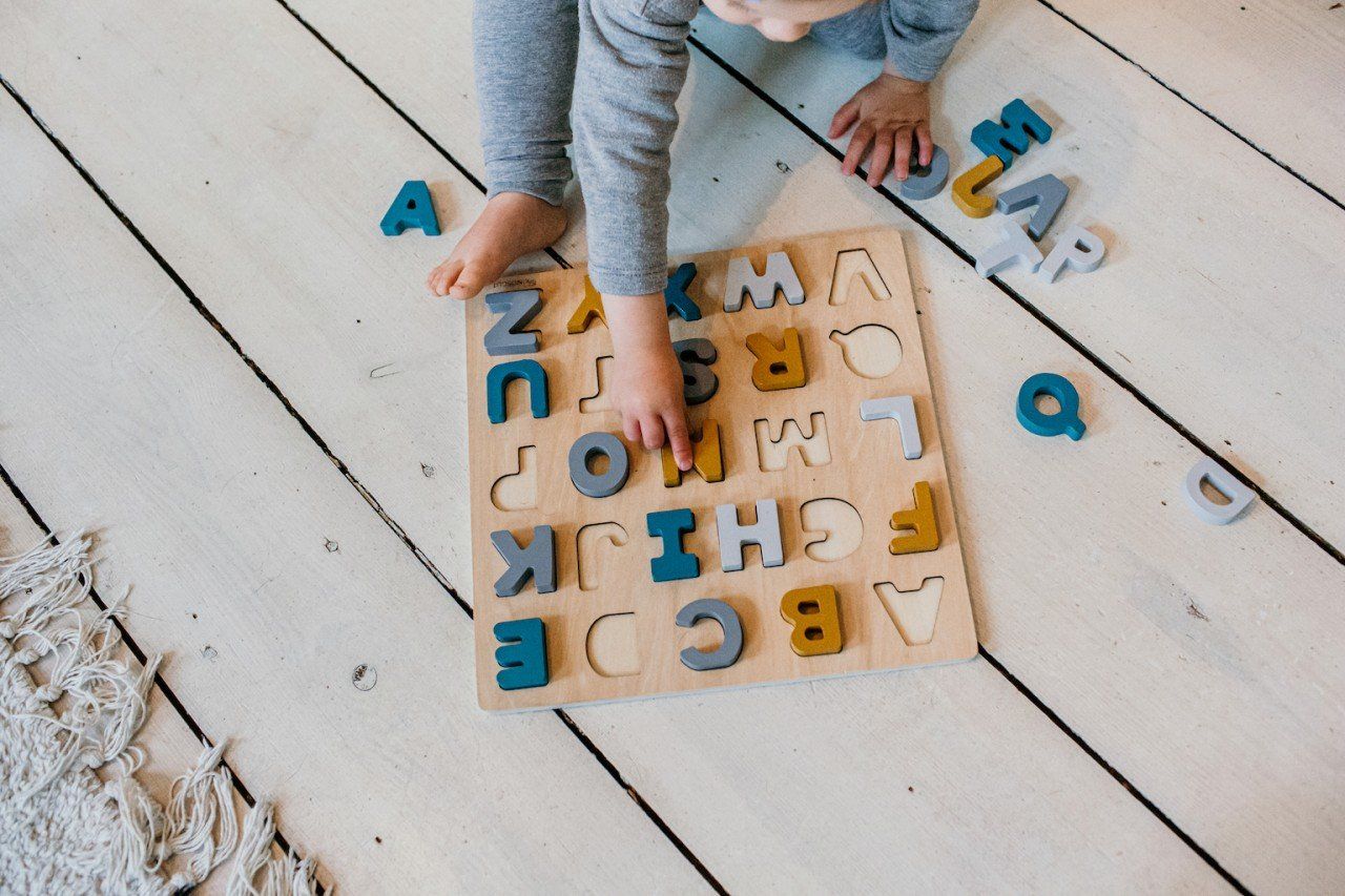Kind spielt mit einem Holzpuzzle. Das Puzzle hat Buchstaben in verschiedenen Farben. Das Kind sitzt auf dem Boden.