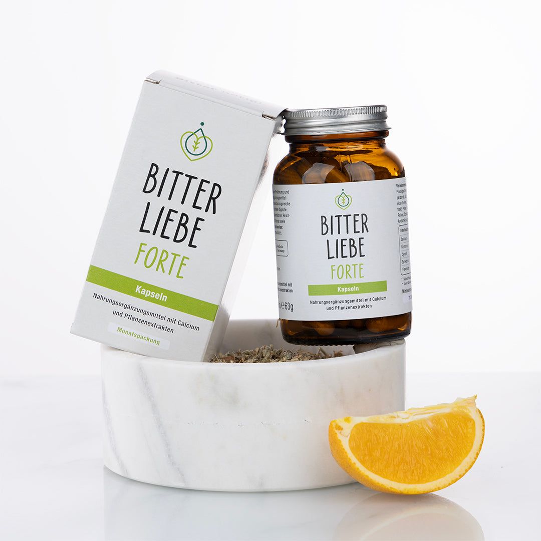 Braune Glasflasche und Schachtel mit "BitterLiebe Forte". Flasche auf weißem Sockel, daneben eine Orangenscheibe.