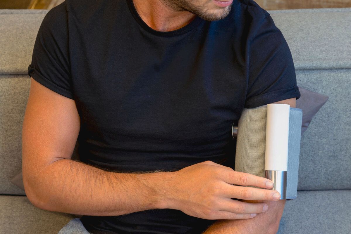 Person mit Blutdruckmessgerät am Arm. Graues Manschette, weißer Zylinder. Mitings-Logo.