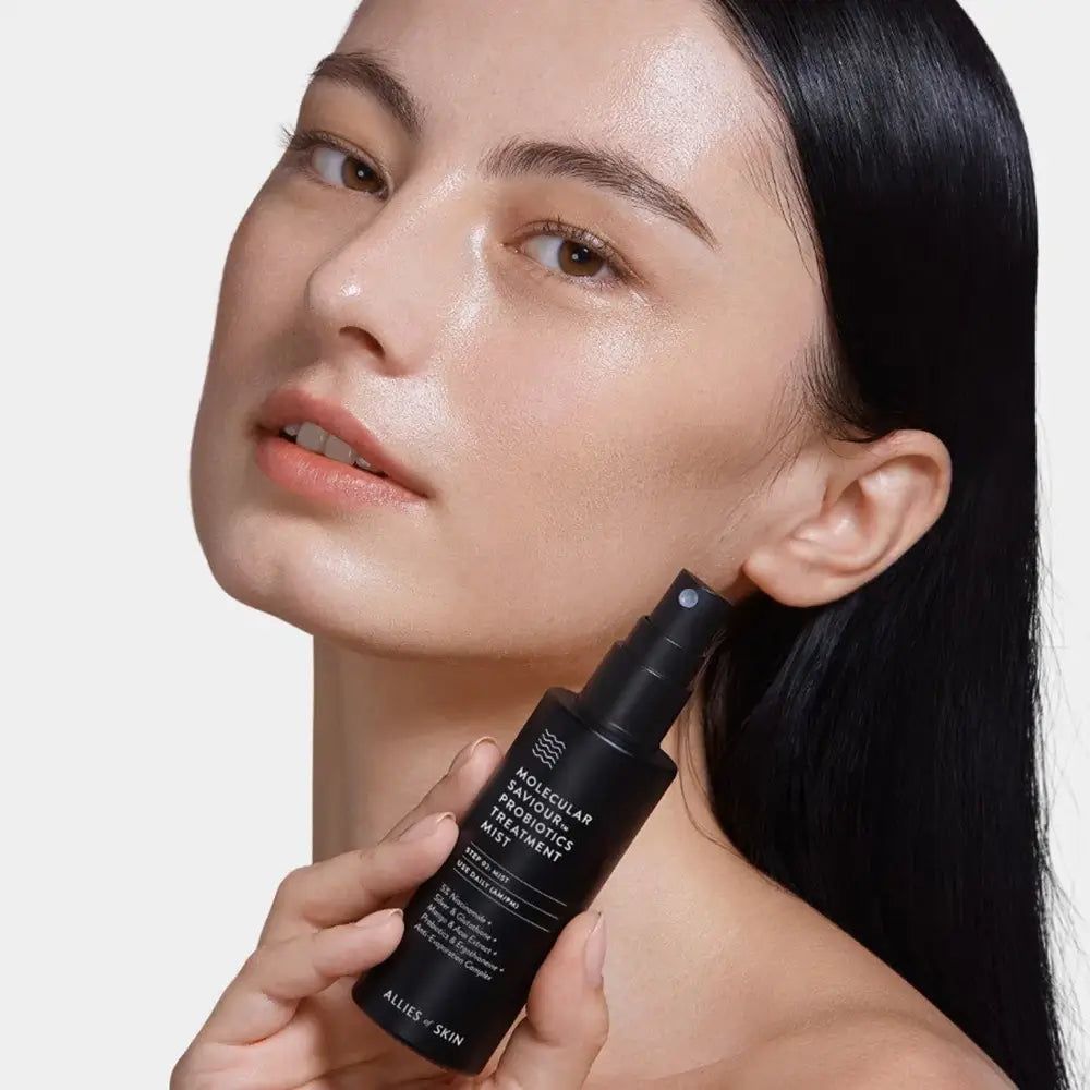 Frau hält eine schwarze Sprühflasche. Auf der Flasche steht: Molecular Saviour Probiotics Treatment Mist. Sichtbar: 5% Niacinamid, Silber, etc.