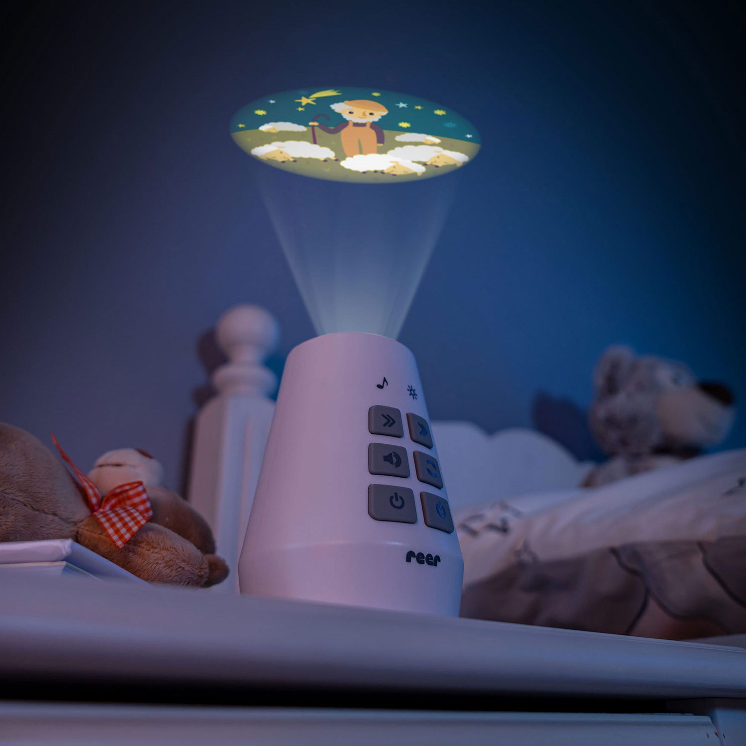 Projektor mit Projektion auf Decke. Teddybär auf Nachttisch. Logo 'reer' auf Projektor.