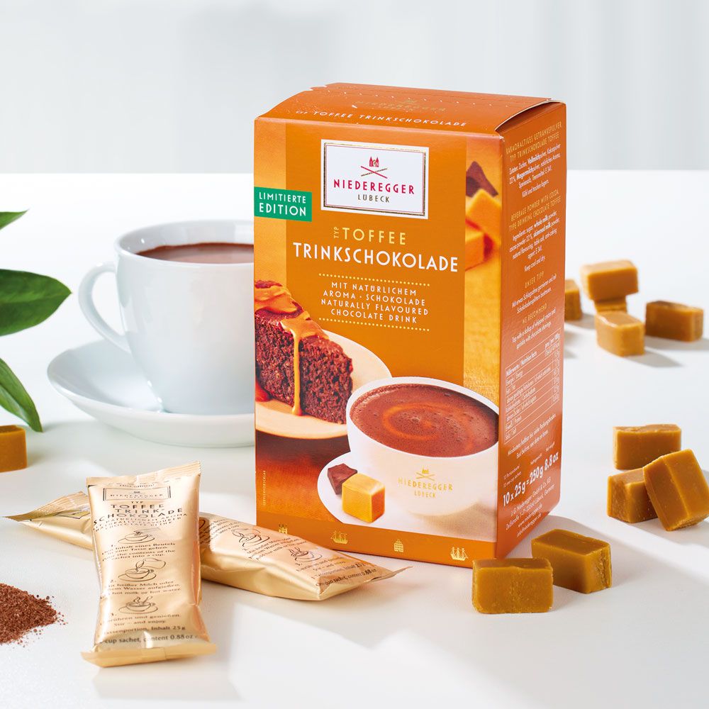 Niederegger Toffee-Trinkschokolade, Verpackung mit Produktabbildung. Einzelne Toffee-Stücke und Trinkschokoladenbeutel daneben.