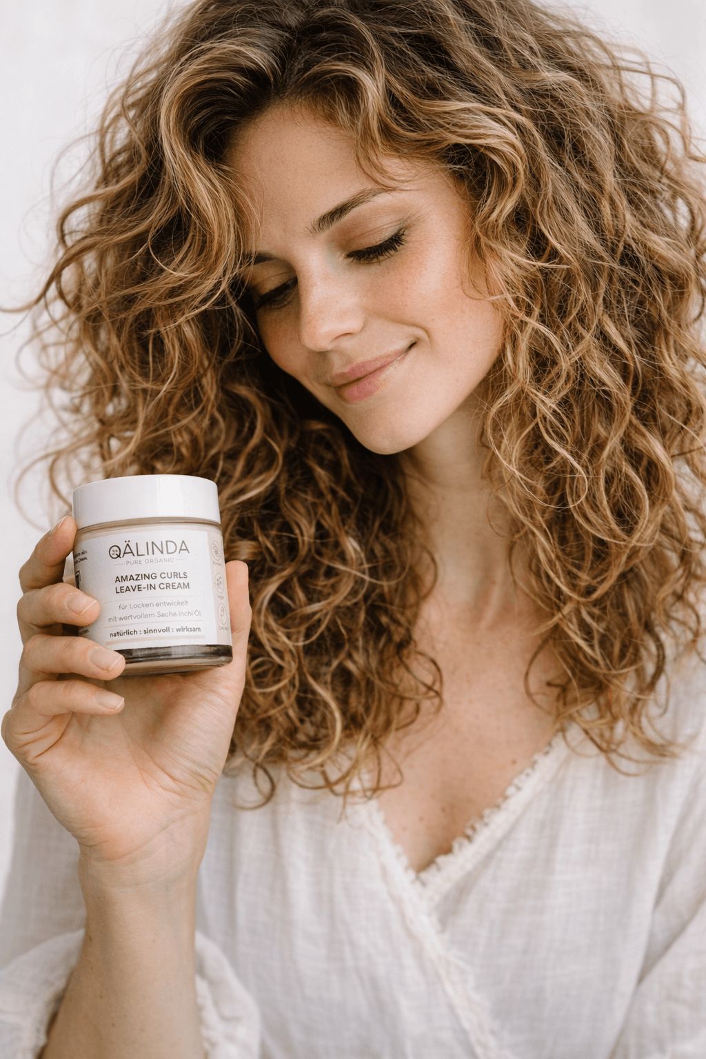 Frau hält einen Tiegel QAELINDA Amazing Curls Leave-In Cream. Frau mit lockigem Haar, helles Oberteil. Produkt in der Hand.