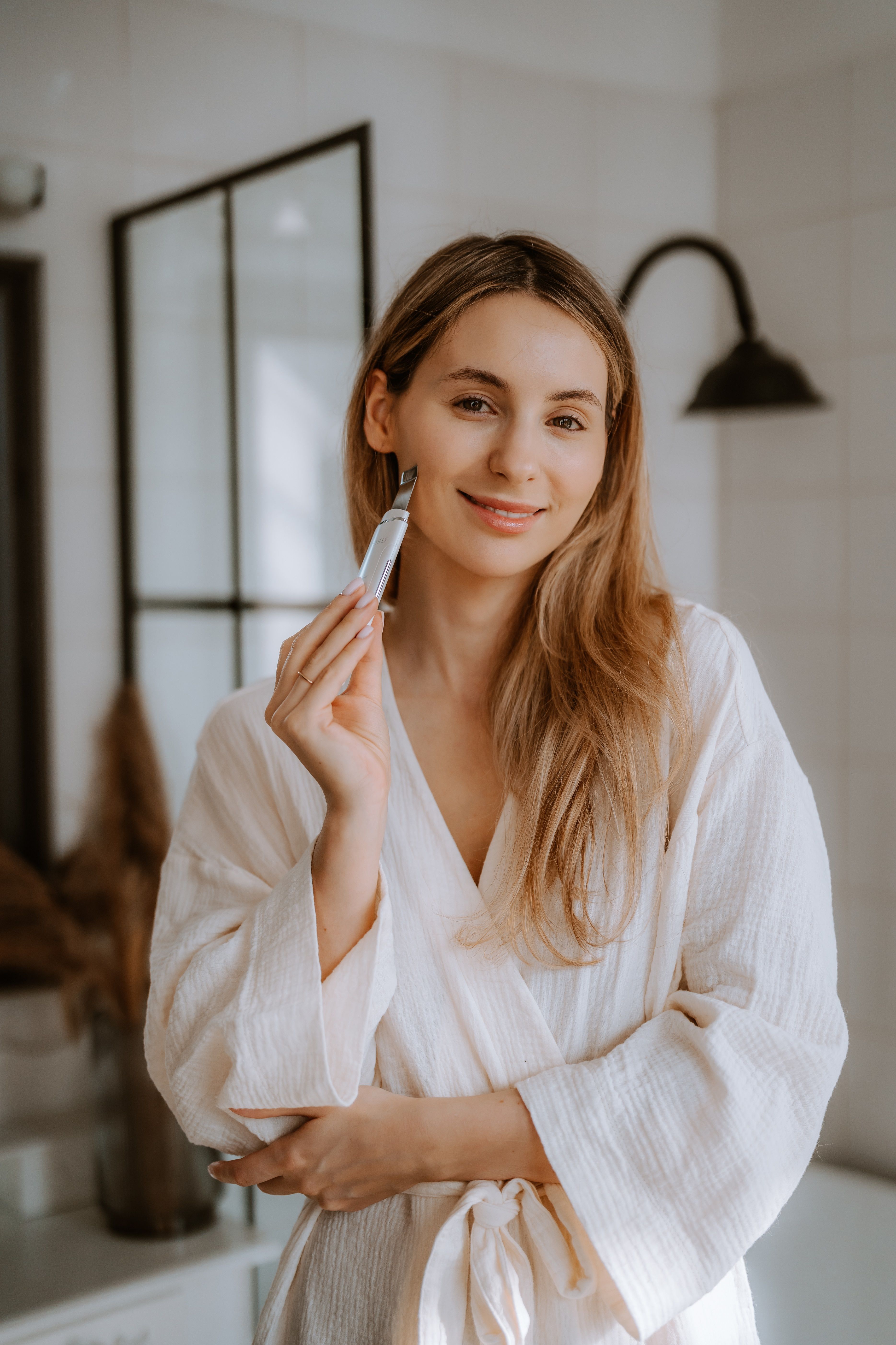 Frau hält ein weißes BEAUTIFLY B-Scrub Young Ultraschallpeelinggerät. Frau trägt einen weißen Bademantel. Badezimmer im Hintergrund.