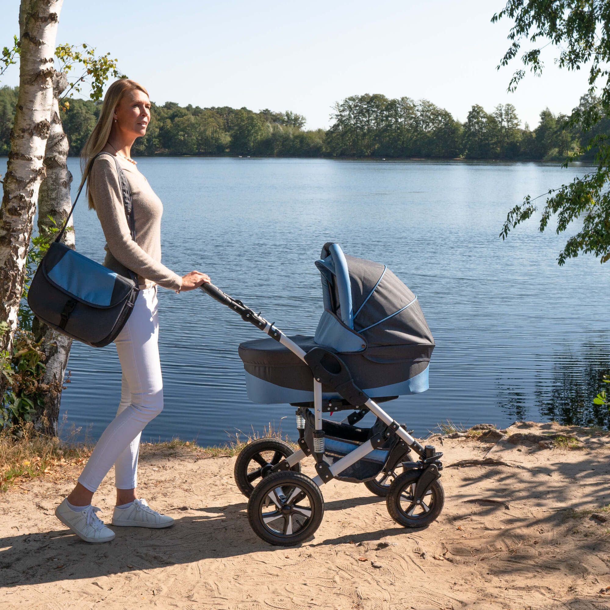 Frau schiebt Kombikinderwagen, grau-blau, am Seeufer. Tasche an der Schulter. Hartgummireifen. Sonniges Wetter.