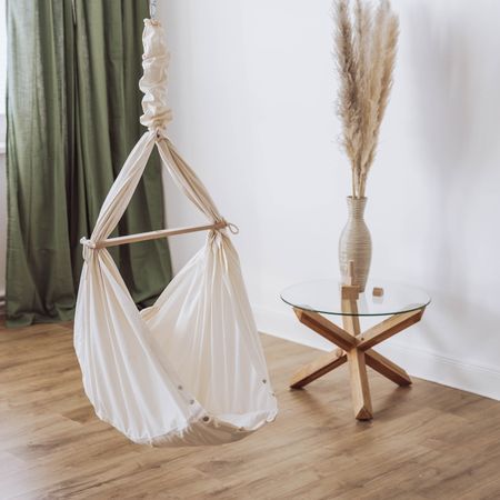 Babyhängematte in beige. Hängematte hängt an Feder. Steht neben Tisch mit Vase und Pampasgras.