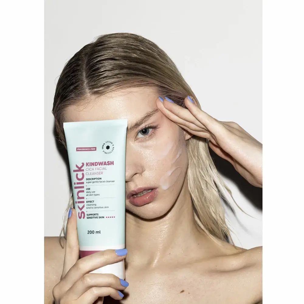 Frau hält Tube vor Gesicht. Aufschrift: Kindwash Cica Facial Cleanser. Gesicht mit Produkt bedeckt. Hellblauer Hintergrund.