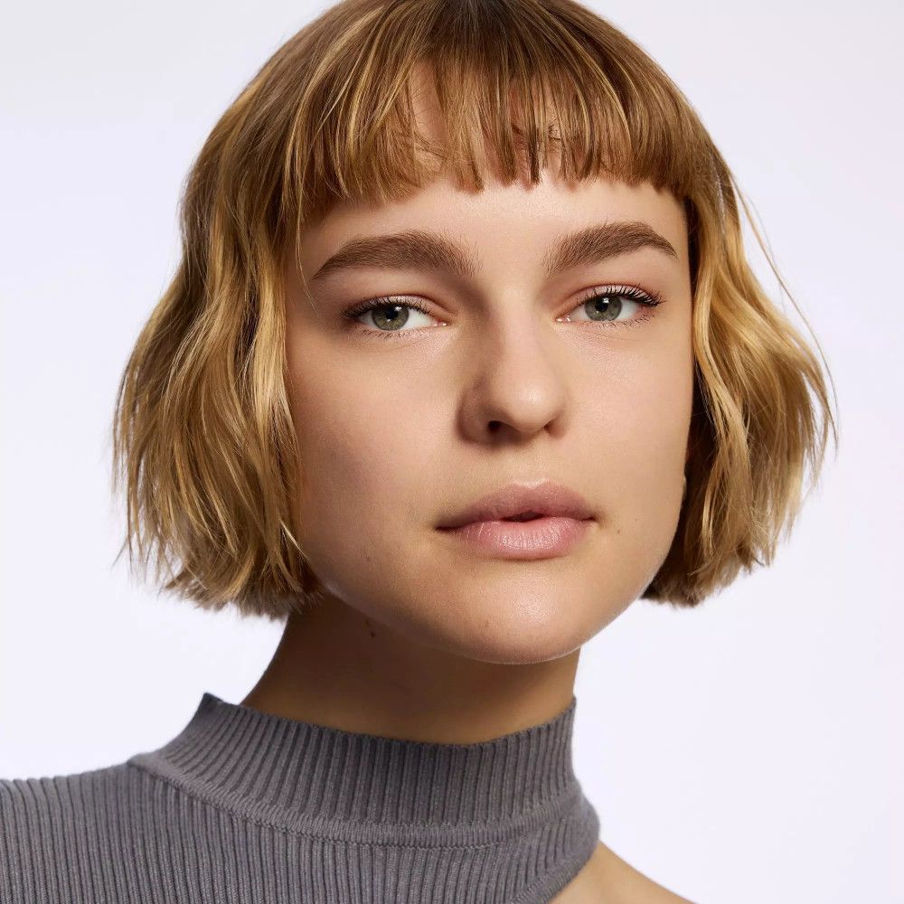 Porträt einer Frau mit kurzen blonden Haaren und grauem Oberteil. Gesichtsausdruck und Hautton sichtbar.